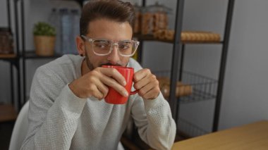 Kapalı bir iş yerinde kırmızı bardaktan kahve yudumlayan genç adam rahat, düşünceli bir atmosfer sunuyor modern ofis ortamında raflar ve bitkilerle..