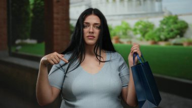 Parkta durmuş, ciddi bir ifadeyle elinde mavi bir alışveriş çantası olan, kıvrımlı, büyük beden ve uzun siyah saçları olan bir kadın..