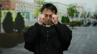 Dini giyinmiş, elleri kelepçeli bir adam şehir caddesinde duruyor, inanç ve baskı arasında çarpıcı bir tezat oluşturuyor, arka planda şehir binaları ve yeşillik var..