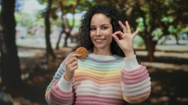 Parkta renkli süveter giyen genç İspanyol kadın ağaçlar ve güneş ışığıyla dolu bir ortamda lezzetli kurabiyelerin tadını çıkarırken iyi bir jest yapıyor..