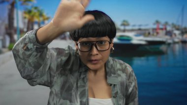 Elleri deniz kıyısında, açık mavi gökyüzünün altında tekneleri olan gözlüklü ve kamuflajlı bir ceket giyen bir kadın, açık havada anlamlı bir an sergiliyor..