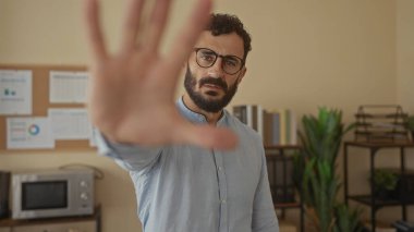 Kapalı ofis ortamında sakallı İspanyol bir adam, çağdaş bir ortamda duraksama ya da tedbir taşıyarak duraksamayı durdurur..