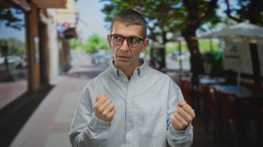 Güneşli bir sokakta ayakta duran gözlüklü ve çizgili gömlekli bir adam, yumruklarını hafifçe sıkmış ve arka planda binalar olan düşünceli görünüyor..