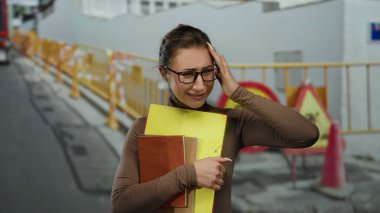 Gözlüklü genç bir kadın, bir inşaat alanında endişeli bir ifadeyle, şehir merkezindeki hayal kırıklıklarını ve evrak işlerini kaydediyor..
