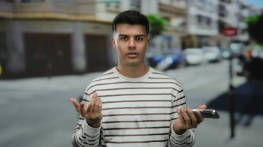 Şehir caddesinde akıllı telefonlu genç bir adam açık havada kargaşa yaratıyor arka planda bulanık araçlar ve binalar olan sıradan çizgili bir gömlek giyiyor..