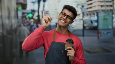 Gözlüklü ve önlüklü genç İspanyol bir adam şehrin işlek caddesinde kahve fincanı tutuyor. Dostane bir ifadeyle yukarı bakıyor. Şehir ortamında coşku ve açıklık olduğunu gösteriyor..