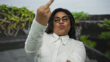Beyaz gömlekli genç Latin kadın, parkta orta parmaklı kaba bir jest yapıyor. Açık havada meydan okuma ve tavır sergiliyor..