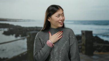 Deniz kenarından hoşlanan uzun saçlı bir kadın kumsala bakıyor, doğal güzelliğini ve huzurlu atmosferini sergiliyor..