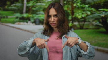 Genç bir kadın, lüks bir parkta iki eliyle jestler yapıyor, rahat bir gömlek giyiyor ve açık havada ciddi bir ifade takınıyor..