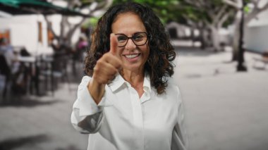 Kıvırcık saçlı olgun bir kadın açık hava restoranının terasında neşeli bir ifadeyle baş parmak kaldırıyor..