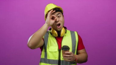 Kasketi ve yansıtıcı yeleği olan genç bir adam canlı pembe arka planda kahve tutarken işaret veriyor..