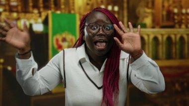 Gözlüklü ve kırmızı örgülü bir kadın kapalı bir kilise ortamında anlamlı bir şekilde konuşur, duygularını aktarır ya da şaşırtır..