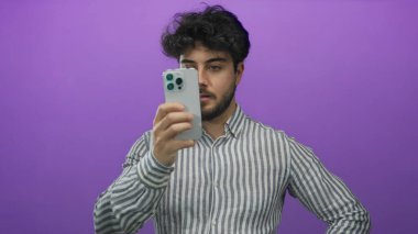 Sakallı genç adam canlı mor bir arkaplan üzerinde çizgili bir gömlek giyerken akıllı telefonu tutuyor, muhtemelen sosyal medya içeriğine mesaj atıyor ya da araştırıyor..