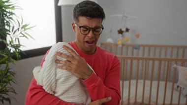 Beyaz bir battaniyeye sarılı gözlük takan genç bir adam rahat bir yatak odasında beşik ve bitkiyle esnerken, ebeveyn bakımı ve ev ortamını gösteriyor..