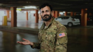 Askeri üniformalı İspanyol adam park yerinde duruyor, kolundaki Amerikan bayrağını gösteriyor, etrafı park halindeki arabalarla çevrili ve loş bir garajla çevrili..