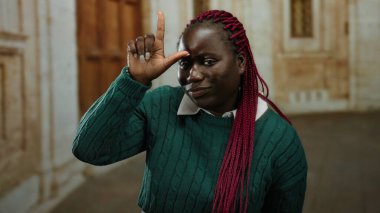 Eski üniversitede kamera karşısında zavallı bir jest yapan kadın neşeli bir ifade ve örgülü saçla eğitim ortamında kendine güven ve mizah gösteriyor..