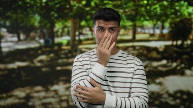 Çizgili tişörtlü İspanyol adam gölgeli ağaçlarla kaplı güneşli bir parkta şaşırmış görünüyor..