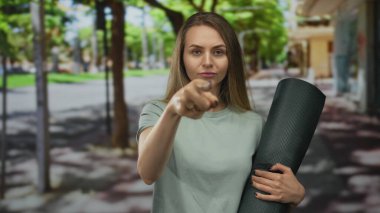 Elinde yoga minderiyle sokakta ağaçlarla el kol gezen ve arka planda bulanık binalar olan bir kadın şehir yaşamını işaret ediyor..