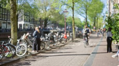 Amsterdam 'ın güneşli bir kanalı boyunca yürüyen ve bisiklete binen, yemyeşil ve caddeleri çevreleyen bisikletli insanların bulanık bir ilkbahar manzarasını görüntülemesi..