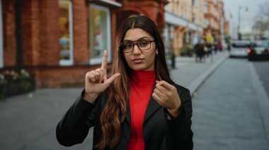 Gözlük takan kadın, şehir sokağında arka planda kırmızı tuğlalı binalarla ezik jestler yapıyor. Dışarıdaki hareketli ortamda kendine güvenen tavırlar sergiliyor..
