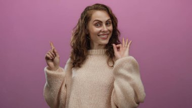 Süveter giyen genç bir kadın pembe bir arka plana işaret ediyor. Yeni stüdyo ortamında pozitiflik ve sadelik taşıyor..