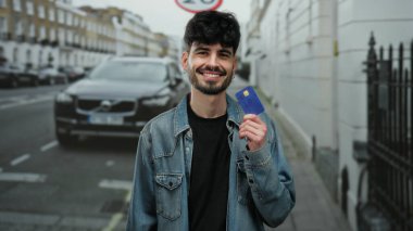 Kentsel sokak ortamında kredi kartıyla gülümseyen, kot ceket giyen, park halindeki arabanın önünde duran, modern ekonomik kolaylığı gösteren genç bir adam..
