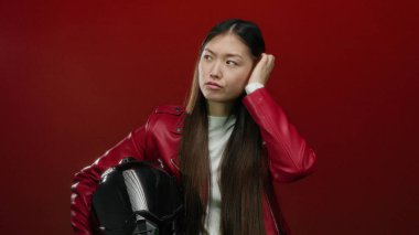 Kırmızı ceketli bir kadın kırmızı arka planda miğfer tutuyor düşünceli ve odaklanmış görünüyor.