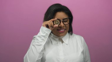 Genç Latin bir kadın pembe arka planda kripto para tutuyor, izole bir duvarda kripto finansmanı sergiliyor..