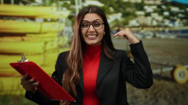 Genç İspanyol kadın deniz kıyısındaki bir yeri işaret ediyor. Elinde açık mavi gökyüzü ve uzak evleri olan güneşli bir arka planda kırmızı bir pano var..