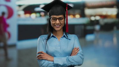 Şapkalı ve cüppeli genç bir kadın, yoğun bir havaalanı terminalinde kollarını kavuşturarak güvenle gülümsüyor, gelecekteki yolculuklar için bir başarı ve istek duygusu simgeliyor..