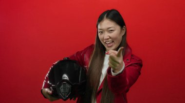 Deri ceket giyen, modern tarzı ve kendine güveni olan, neşeli bir miğferi olan, kırmızı arka planda duran bir kadını işaret ediyor.