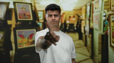Beyaz tişörtlü genç adam, sanat galerisinde parmağı olmayan, kapalı alanda renkli resimlerle çevrili, yüzünde ciddi bir ifadeyle..