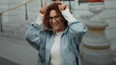 Woman brunette middle-aged smiling making horns gesture with fingers wearing glasses on a city street outdoor with blurred background showing urban lifestyle