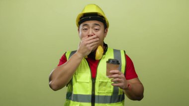 Yorgun Asyalı inşaat işçisi sarı arka planda kahve tutarak esniyor güvenlik ekipmanları ve miğfer takıyor iş sırasında yorgunluk hissi veriyor..
