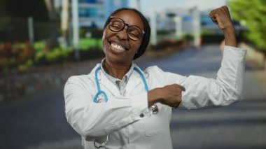 Gözlüklü ve steteskoplu kendine güvenen kadın doktor şehir merkezinde kollarını esnetiyor ve gururlu bir gülümsemeyle şehir ortamında güç ve profesyonellik öneriyor.