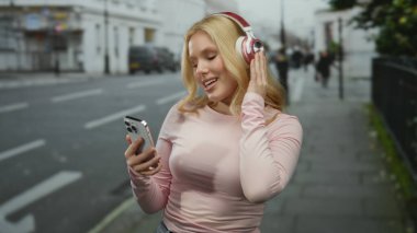Kulaklıklı müzik dinleyen kadın, şehir caddesinde yürürken akıllı telefon tutuyor ve dışarıda günün tadını çıkarıyor..