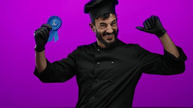 Siyah şef üniformalı İspanyol adam, mor arka plana karşı birinci sınıf mavi kurdeleyle kutlama yapıyor, neşe ve başarıyı ifade ediyor..
