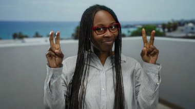Kıvırcık saçlı bir kadın deniz kenarında gezinirken tırnak işareti yapıyor. Plaj zemininde gözlük takıyor ve bulutlu bir gökyüzünün altında rahat bir ifade takınıyor..