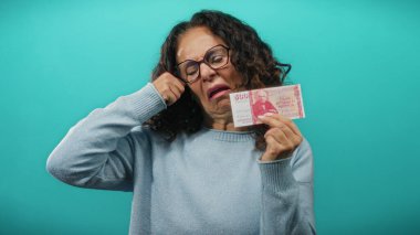 Turkuaz arka planda mavi kazaklı orta yaşlı bir kadın elinde İzlanda Krona banknotları tutarken maddi sıkıntıları yansıtan üzgün bir ifade takınıyor..