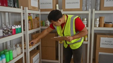 Bağış merkezinde çalışan genç bir adam yansıtıcı yelek giyiyor ve düzgün düzenlenmiş yiyecek eşyaları ve kutular arasında bir pano tutuyor..