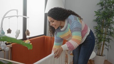 Sıcak bir yatak odasında, renkli bir süveter ve bebek arabasıyla, beşikte gülümseyen bir kadın, sıcak, sıcak bir ortam yaratıyor, ev içinde, şefkatli ve neşeli bir atmosferi vurguluyor..