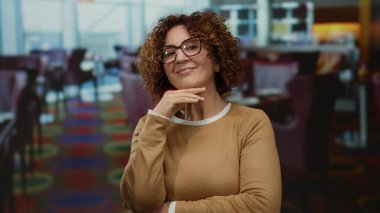 Lokanta ortamında parlak bir dekorla gülümseyen bir kadın gözlük ve kazakla düşünceli bir şekilde poz verirken hareketli bir yemek alanında huzurlu bir güven anı yakalıyor..