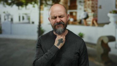 Sakallı ve dövmeli bir adam, canlı bir şehir ortamında boğazını tutarak acı çekiyor..