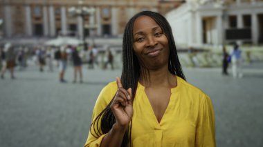 Sarı gömlekli, saçları örgülü bir kadın arkada bulanık yayalar ve tarihi mimariyle çevrili olarak Vatikan meydanında duruyor..