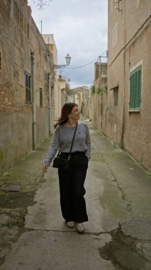 Bir kadın, Mallorca 'nın manzaralı eski bir şehir sokağında mutlu bir şekilde geziniyor, kırsal Akdeniz mimarisi ve enerjik enerjisiyle çevrili, bu tarihi çevrenin cazibesini kucaklıyor..
