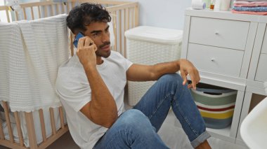 Yatak odası zemininde sakallı genç bir adam, rahat bir evde telefonda konuşuyor, günlük kıyafetler giyiyor, bebek mobilyası ve çamaşır sepetiyle çevrili..