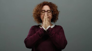 Orta yaşlı, kahverengi saçlı, kestane rengi süveter giyen bir kadın, gri bir arka plana karşı gözleri kapalı meditasyon yapıyor, huzur ve konsantrasyon yayıyor..