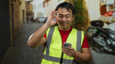 Güneşli bir günde şehir sokaklarında akıllı telefon kullanırken gülümseyen yüksek görüşlü genç adam, şehir yaşamında modern iletişim ve teknoloji kullanımını sergiliyor.