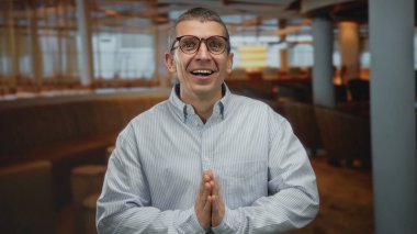 Şaşırmış bir adam restoranda duygularını yüzyüze ifade ediyor, gözlük takıyor ve arka planda bulanık koltuklar ve ışıklar olan çizgili bir gömlek giyiyor..