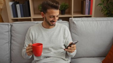 Kanepede dinlenen genç adam, elinde kırmızı bir kupayla telefonuna bakıp gülümsüyor, şık dairesinde rahat bir oturma odası yaratıyor..